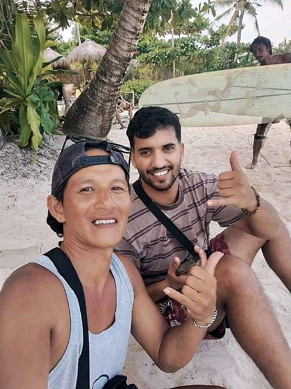 Un moniteur et un élève font le shaka et le pouce levé sur le sable, sous un palmier