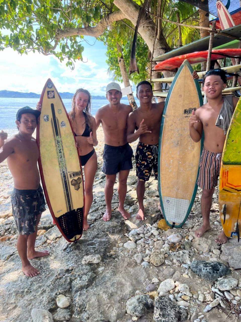 Cinq personnes souriantes avec des planches de surf sur une plage rocheuse, sous les arbres