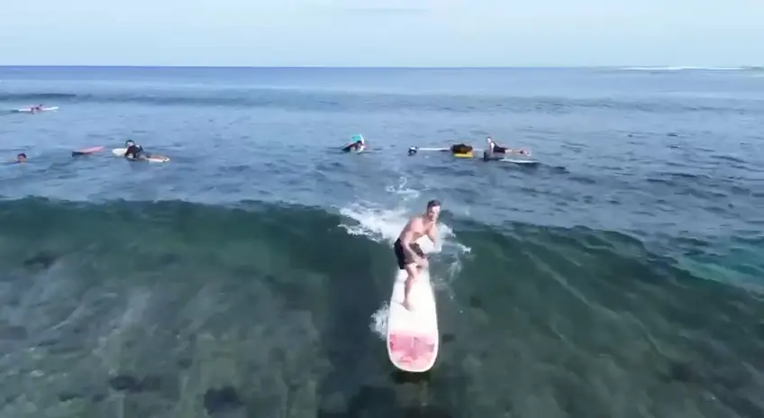Un élève debout sur une planche rose sur une vague claire, filmé par drone, d'autres rament derrière