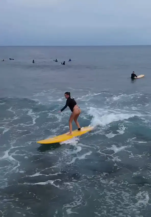 Une élève sur une planche jaune se lève et surfe la vague, filmée par drone